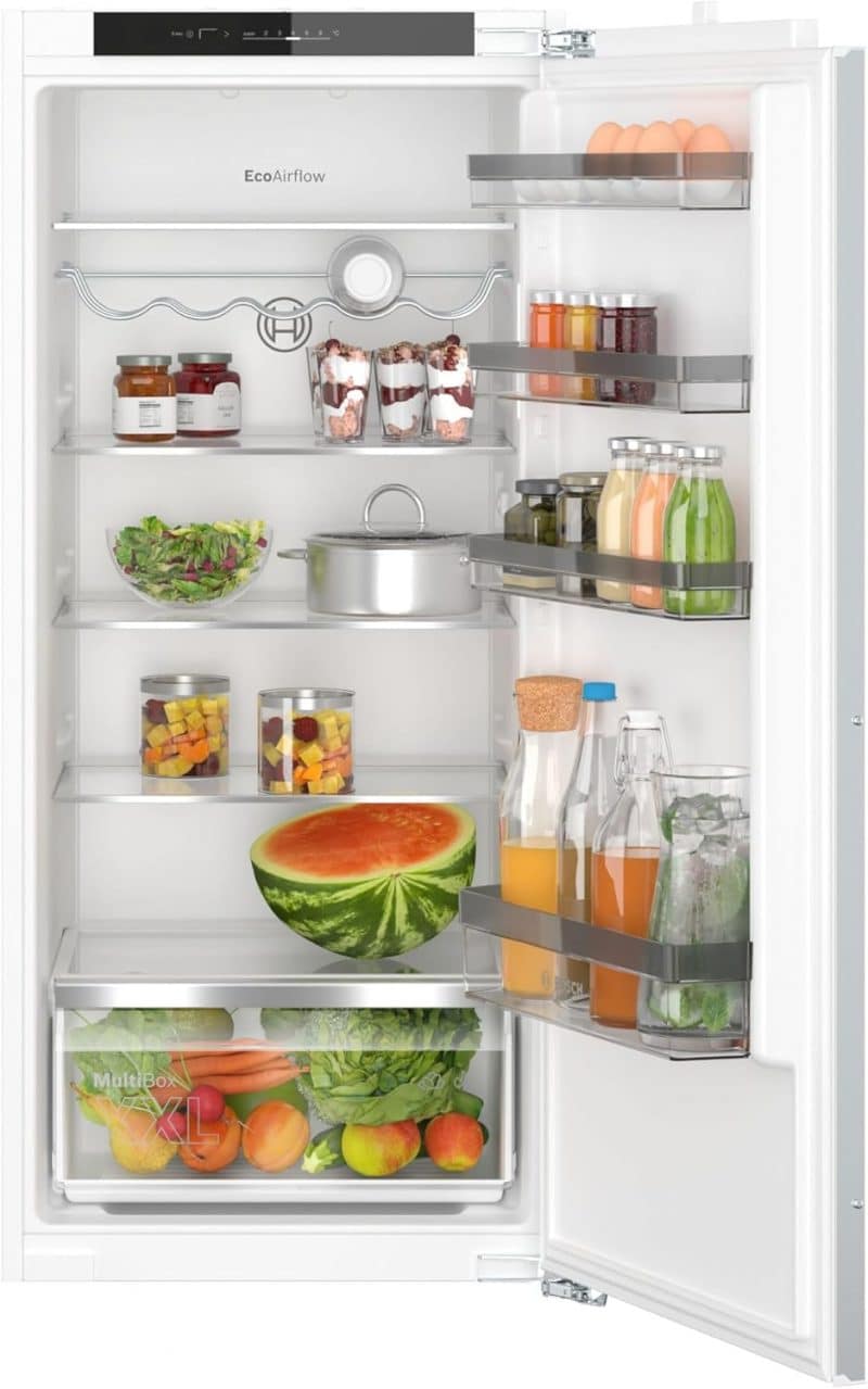 Réfrigérateur encastrable ouvert, rempli de différents aliments frais dont un grand quartier de pastèque, des bouteilles de jus, des bocaux de légumes et un saladier, ainsi que divers fruits et légumes dans le tiroir inférieur. 