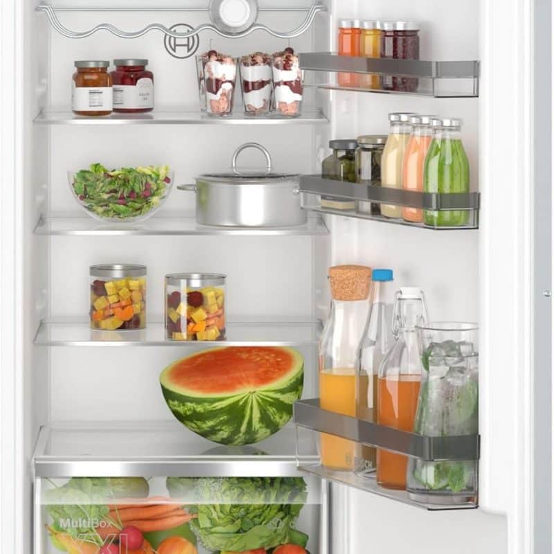 Réfrigérateur encastrable ouvert, rempli de différents aliments frais dont un grand quartier de pastèque, des bouteilles de jus, des bocaux de légumes et un saladier, ainsi que divers fruits et légumes dans le tiroir inférieur. 